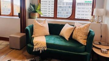 A room with a green sofa, a black and white rug, a potted plant, and a view of buildings through large windows.