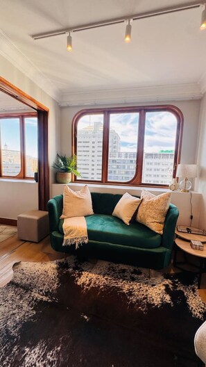 Ein Zimmer mit einem grünen Sofa, einem schwarz-weißen Teppich, einer Topfpflanze und Blick auf Gebäude durch große Fenster.