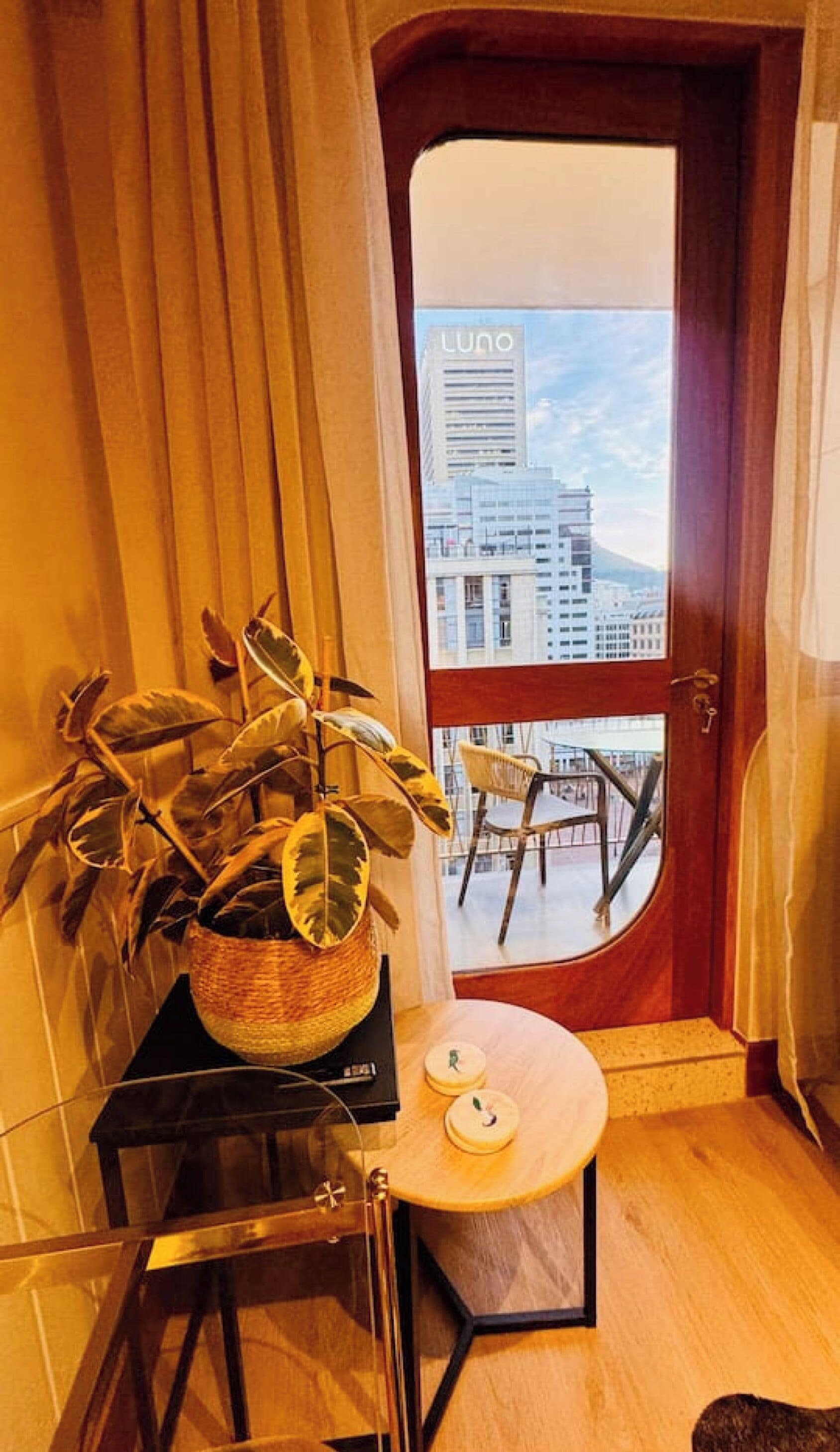 Una habitación con piso de madera, una planta en maceta, una pequeña mesa con dos objetos decorativos, una silla y una puerta que da a un balcón exterior con vista a un edificio identificado como 'Luno'.