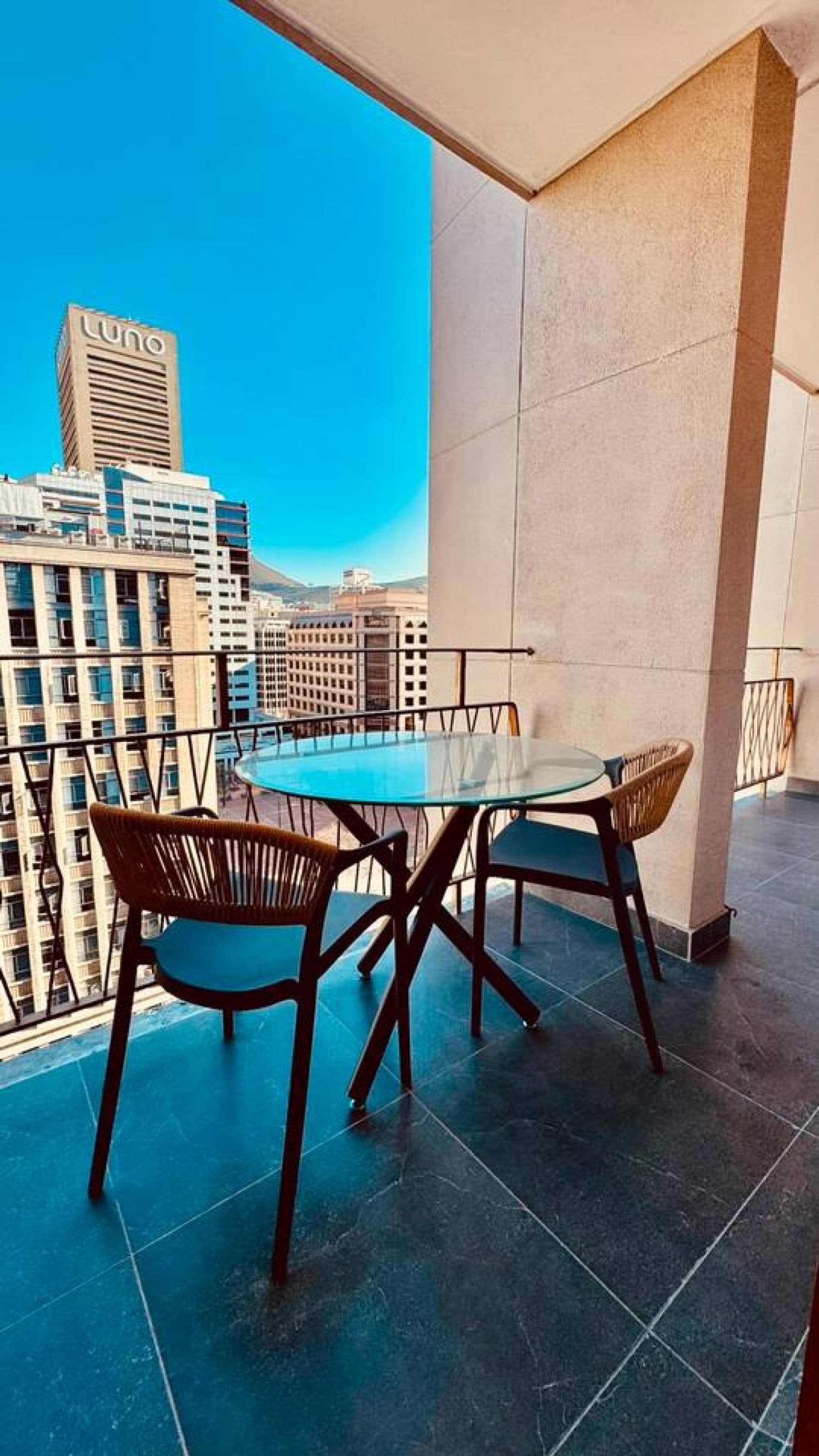 Un balcón con mesa y sillas de vidrio, con vista a un paisaje urbano de edificios altos.