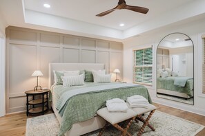 A bedroom with a large bed, two bedside tables with lamps, a ceiling fan, a large mirror, and a window with blinds. - Lowcountry Landing! 3-story townhouse in Old Town's Promenade (Bluffton)