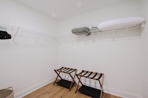 Una habitación pequeña y limpia, con pared blanca, suelo de madera, un tendedero de metal y un estante blanco con mantas dobladas.