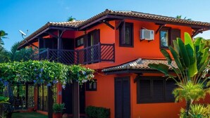 A two-story house with a red exterior, a balcony, and a garden with tropical plants. - Vrbo Property (Acu da Torre (Praia do Forte))
