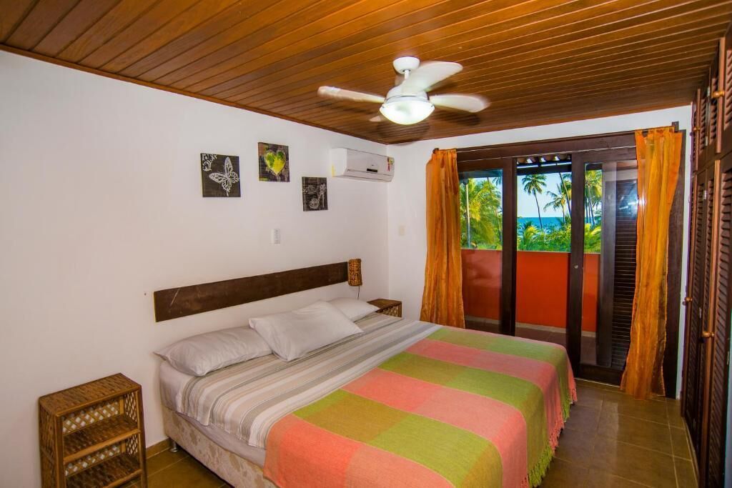 Une chambre à coucher avec un lit, un ventilateur de plafond, une fenêtre avec des rideaux et une vue sur la verdure à l’extérieur.