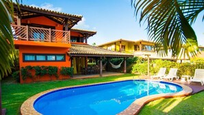 A swimming pool with a tiled edge, surrounded by a well-maintained lawn and a building with a balcony. - Vrbo Property (Acu da Torre (Praia do Forte))