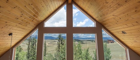 A room with a sloped wooden ceiling and large windows offering a view of a mountainous landscape.