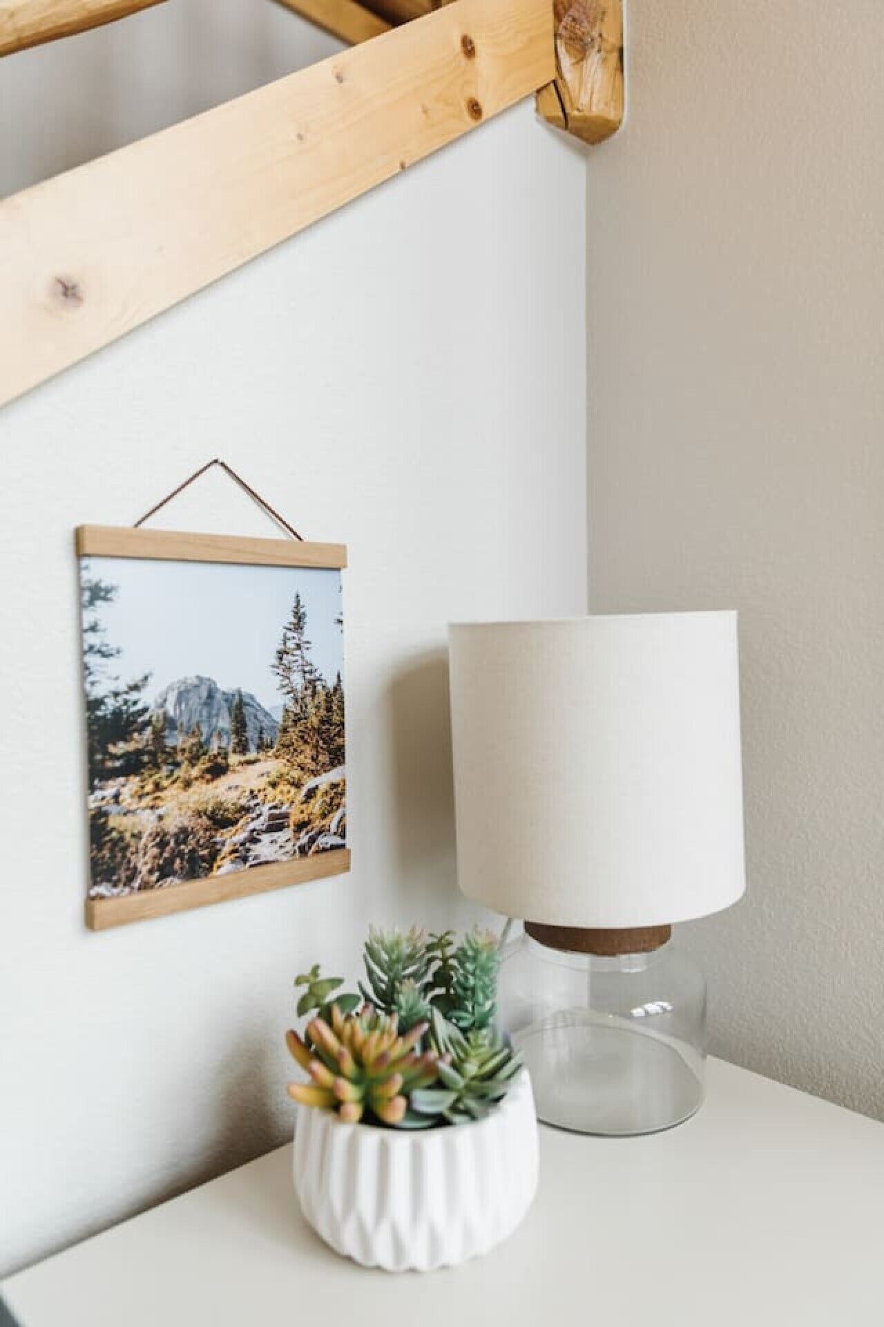 A wall-mounted picture of a mountainous landscape, a white lamp, and a small potted plant on a white surface.