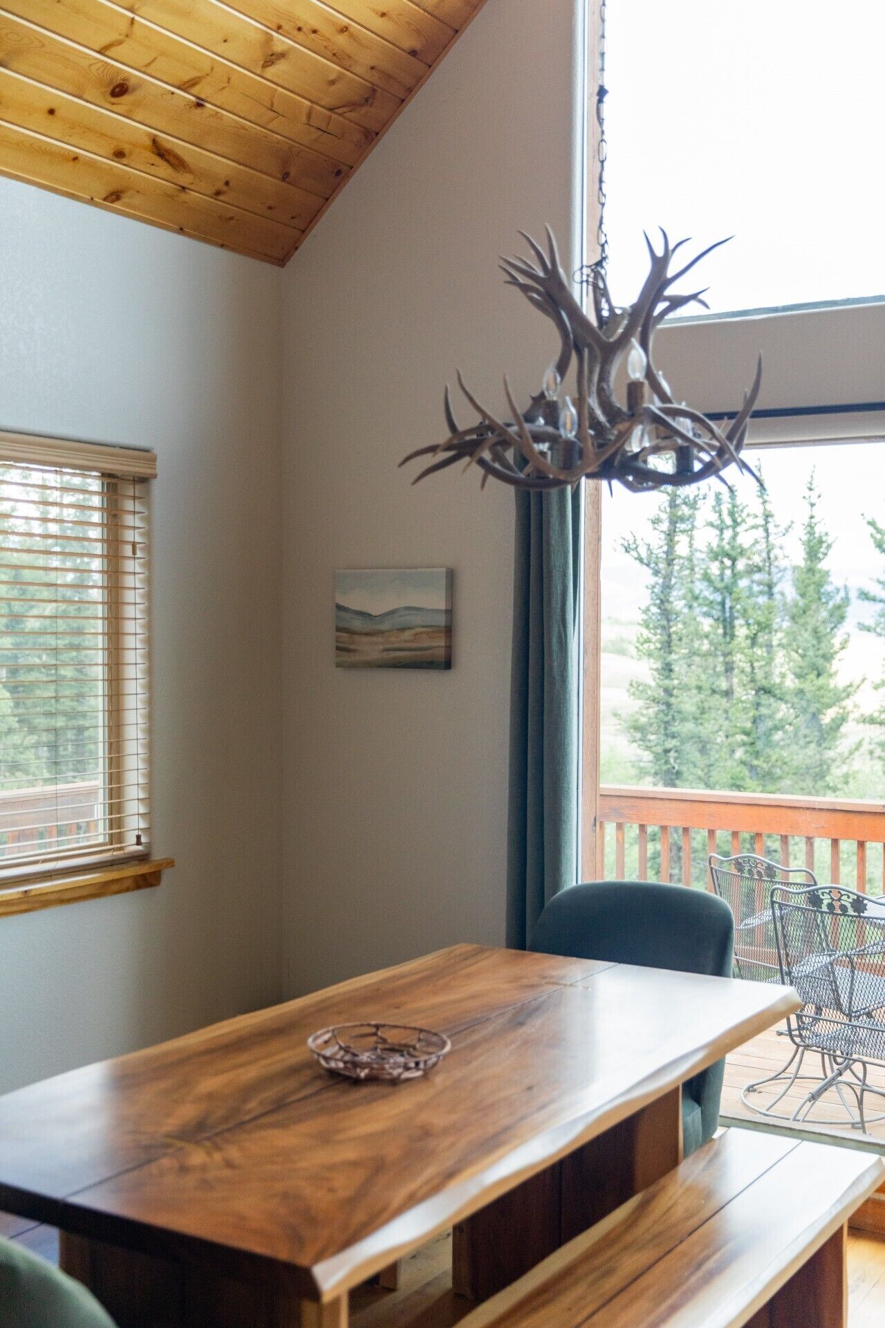 A wooden dining table with benches, a chandelier with antlers, a painting, and a view of trees through a window.