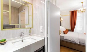 Habitación de hotel con un espejo grande, un lavamanos blanco, una mesita de noche con una planta, una cama con ropa de cama blanca y un cojín marrón, y una araña.
