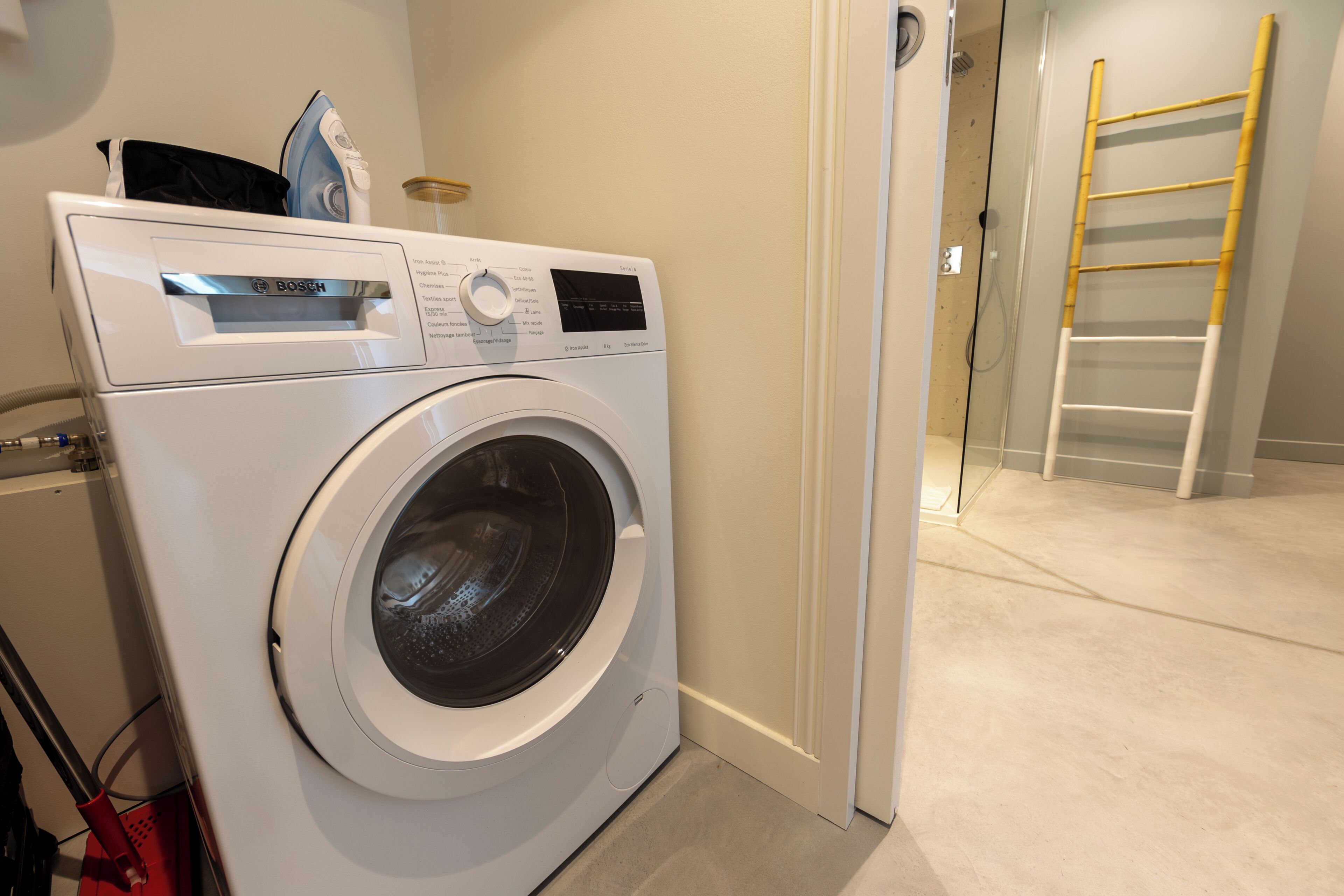A front-loading washing machine in a laundry room with a wooden ladder and a door leading to another room.