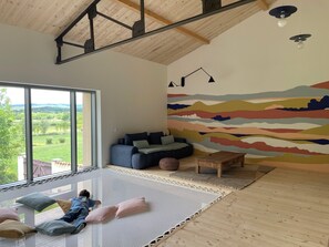 A spacious living room with a large window, a colorful abstract mural, a blue sofa, and a child playing on a mat. - Vacation rental in Haute-Loire with a pool and views of the countryside—perfect for relaxing (Paulhaguet)