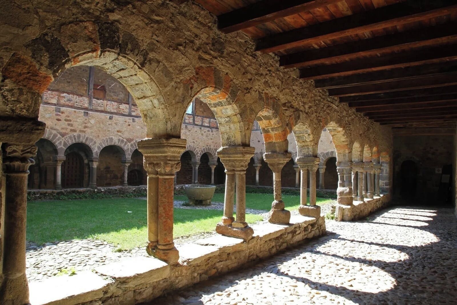 Un antiguo patio de piedra con arcos y columnas, un muro de piedra y una zona con césped.