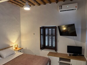 A bedroom with a bed, bedside lamp, TV, air conditioning unit, and a window with blinds. - SimpleGreece Skyros Traditional Holiday Home with Balcony, Wi-Fi & Air Conditioning (Σκύρος)