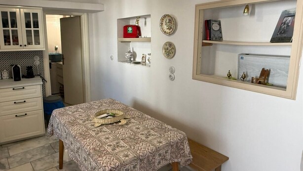 A room with a patterned table, a small bench, a cabinet with glass doors, and a wall-mounted shelf with decorative items. - SimpleGreece Skyros Traditional Holiday Home with Balcony, Wi-Fi & Air Conditioning (Σκύρος)