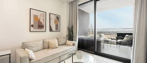 A modern living room with a sofa, a glass coffee table, and a large window offering a view of a balcony with outdoor furniture.