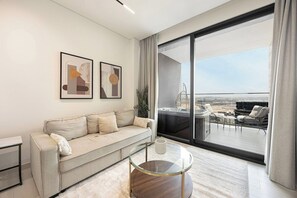A modern living room with a sofa, a glass coffee table, and a large window offering a view of a balcony with outdoor furniture. - Private Jacuzzi / Canal Views / Pool & Gym (Dubai)