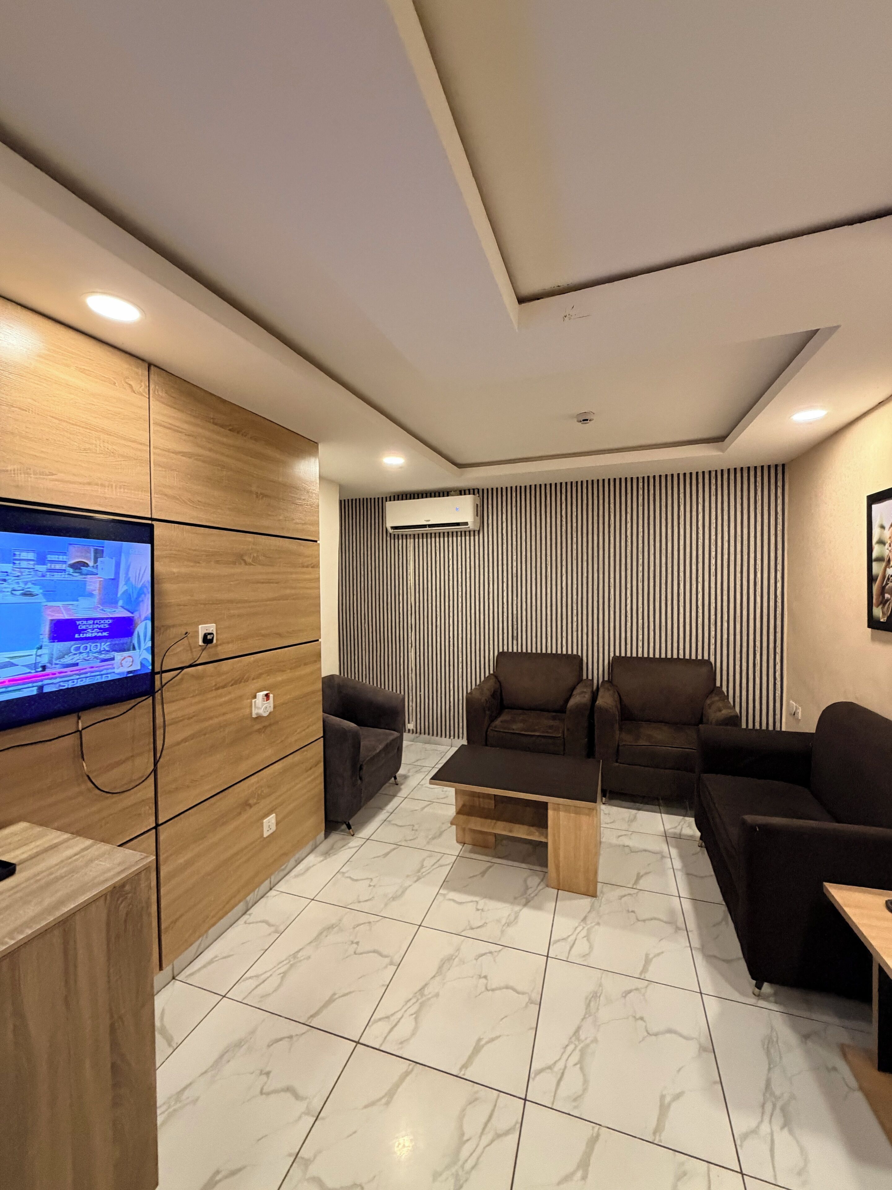A modern living room with a flat-screen TV, dark sofas, a wooden cabinet, and a striped wall.