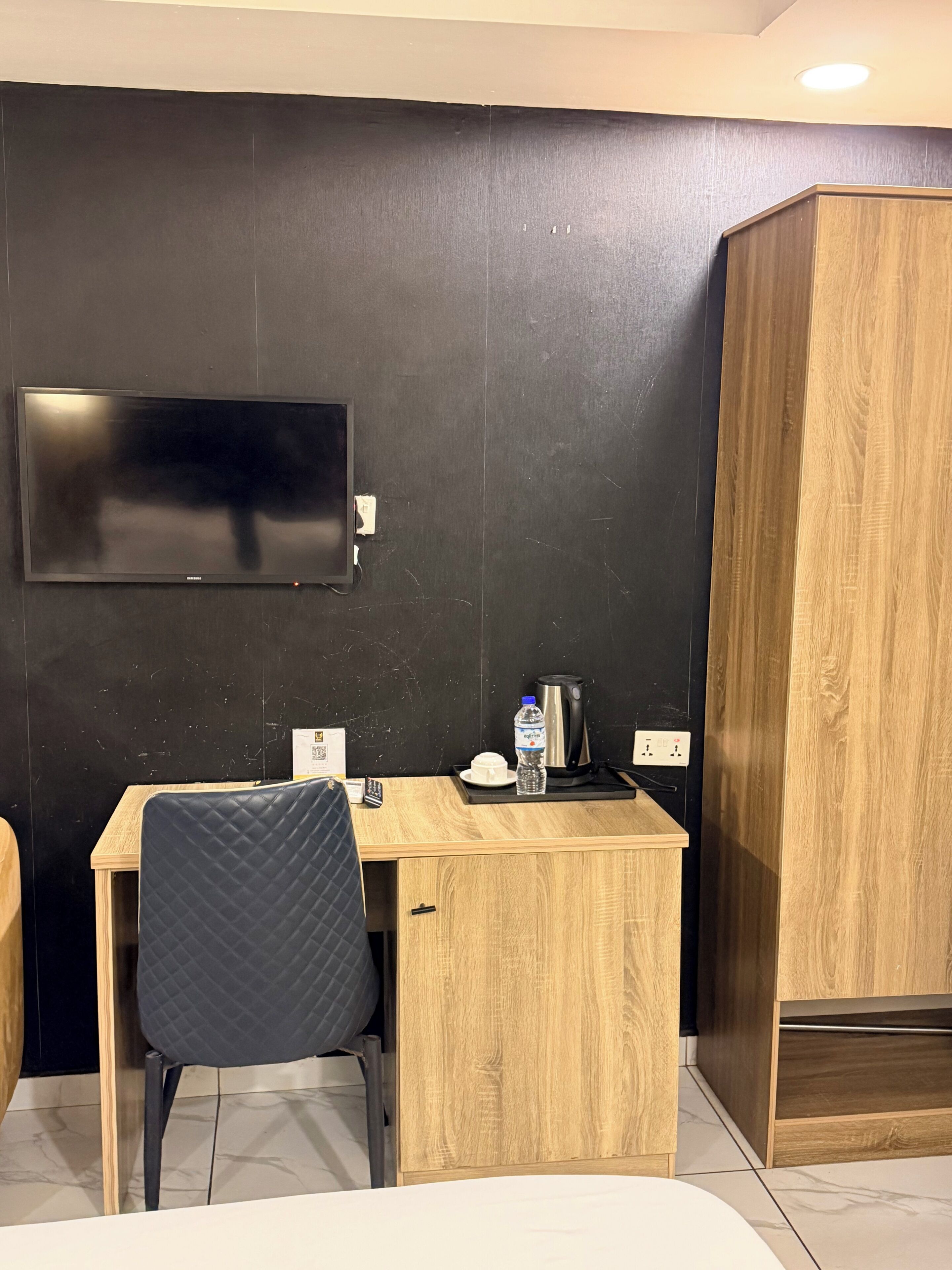 Habitación de hotel con una televisión de pantalla plana montada en la pared, un escritorio de madera con una silla, una botella de agua, un hervidor y una mesita con un plato y una taza.