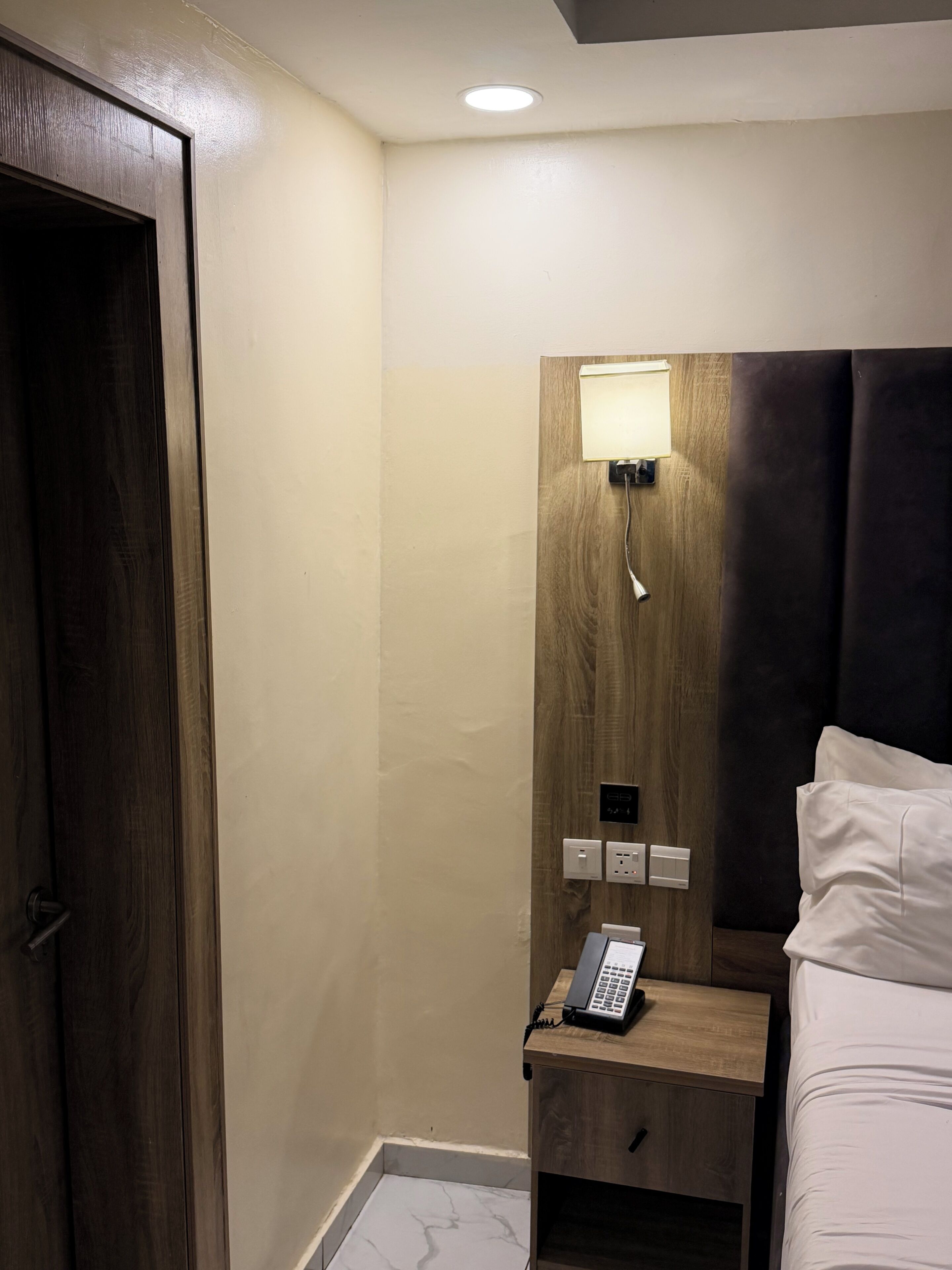 A hotel room with a wooden headboard, a bedside table with a lamp, a phone, and a bed with white linens.