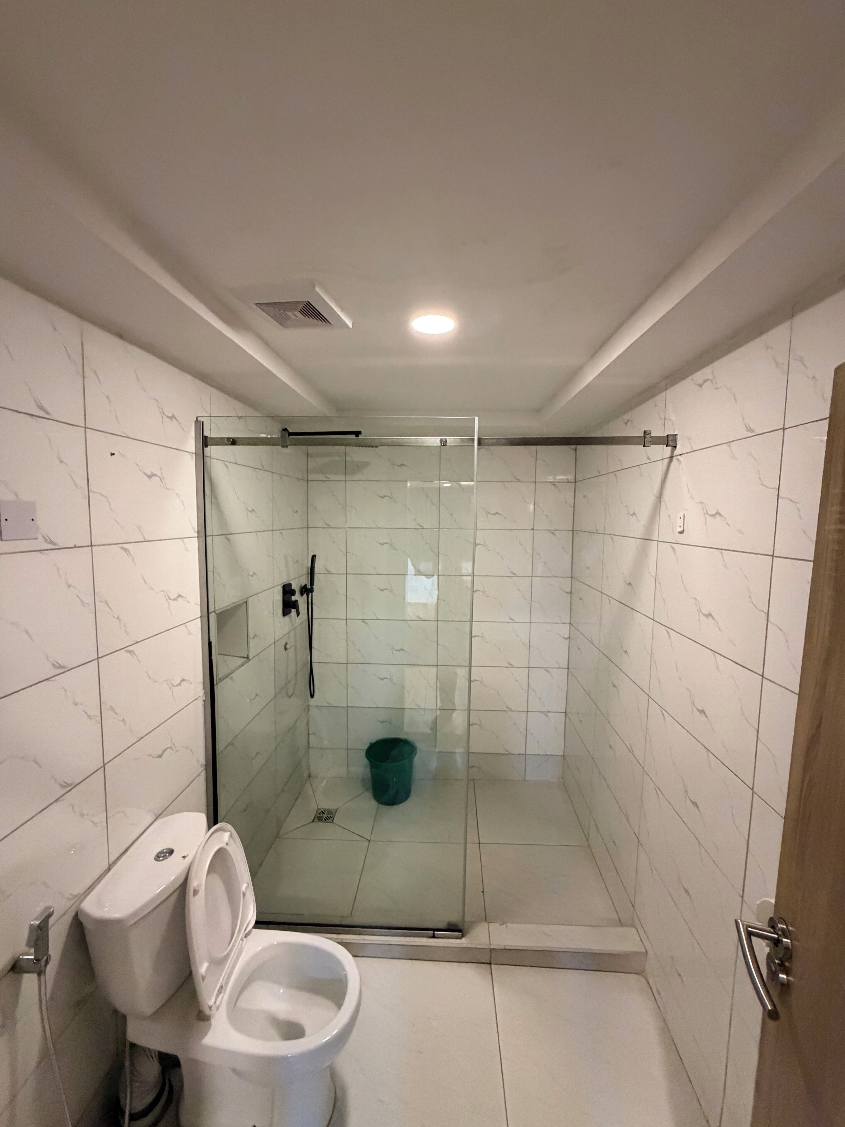 Un baño con ducha envuelta en vidrio, inodoro y un balde.