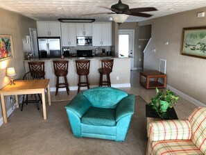 Een zithoek met een blauwgroene fauteuil, een houten salontafel, een gestreepte bank en een keuken op de achtergrond.