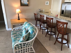 A room with a wicker chair, a wooden table with a lamp, and a kitchen area with a refrigerator and bar stools. - Water Front Delight - Private Beach and Dock, Wi-fi, Pet Friendly, 3 Master BR's (Norfolk)