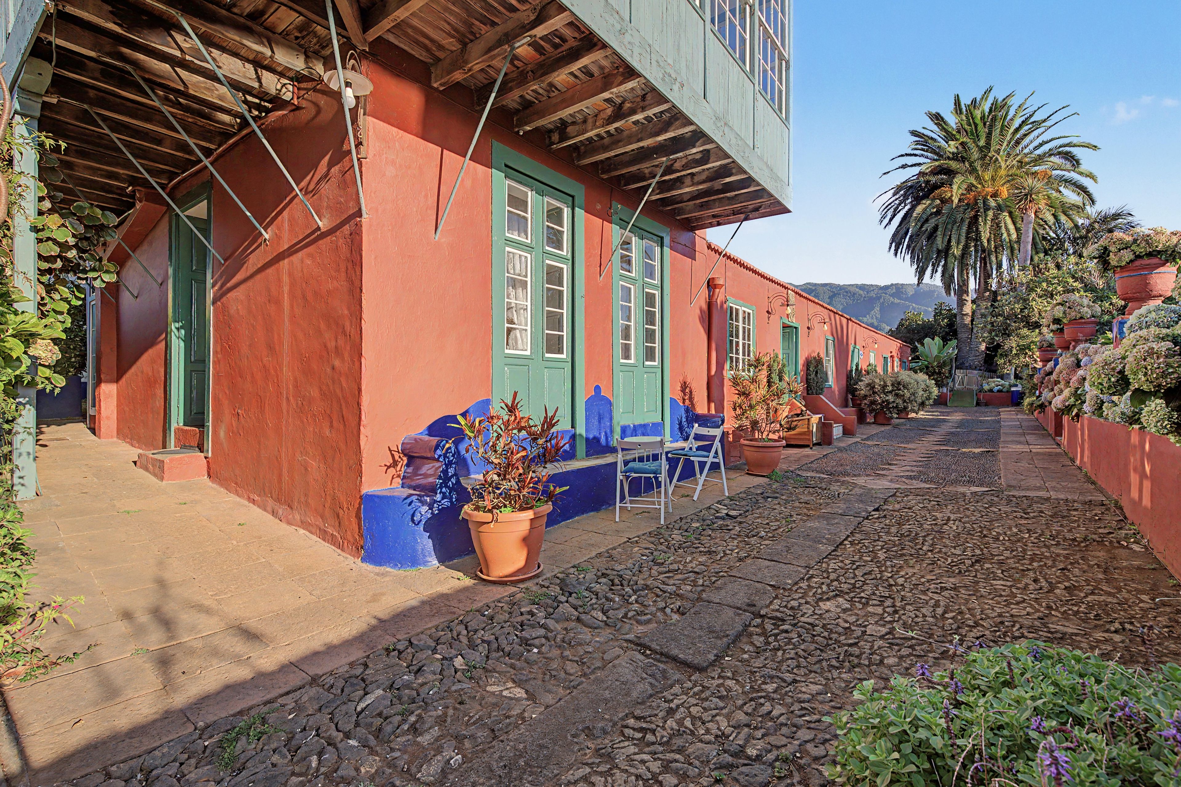 Una estrecha calle adoquinada con edificios pintados de rojo y azul, macetas con plantas y un cielo despejado.