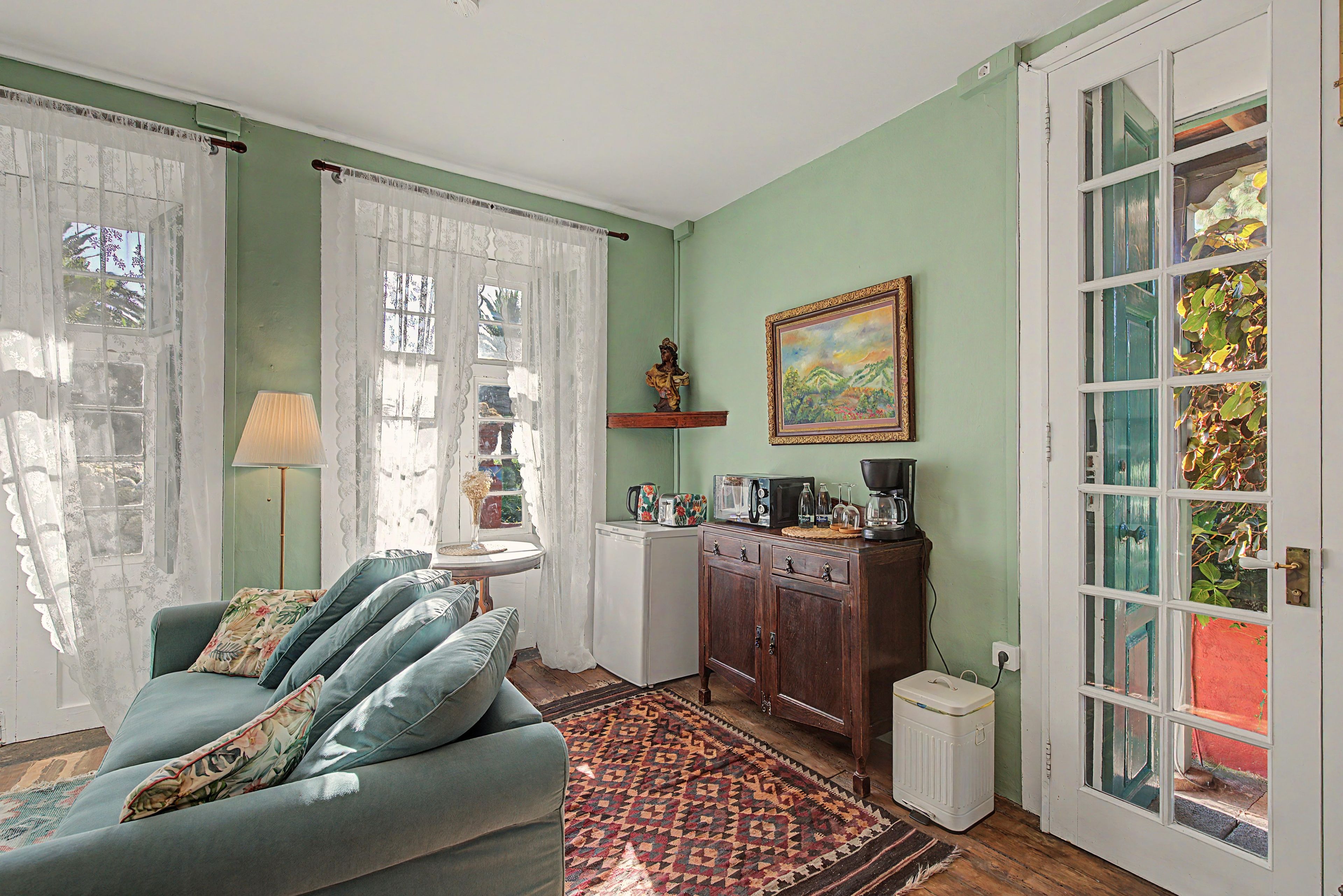 A room with a green wall, a sofa with floral cushions, a small table, a cabinet with kitchen appliances, and a door with glass panels.