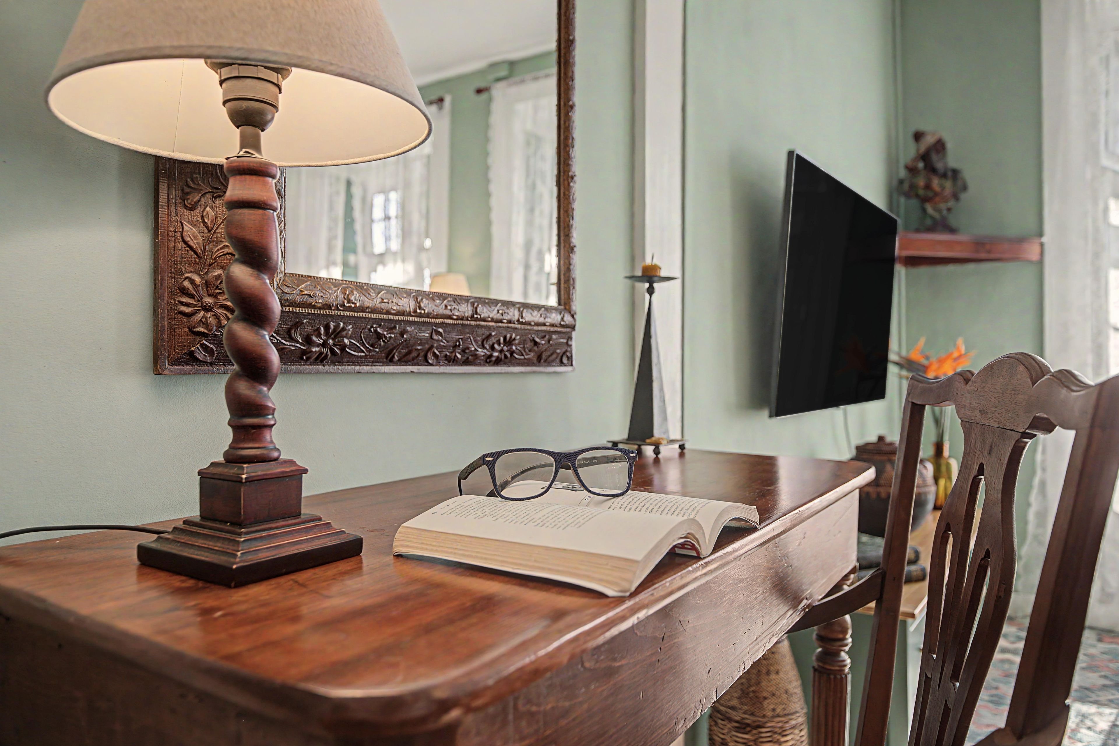 A wooden table with a lamp, a book, glasses, and a mirror.