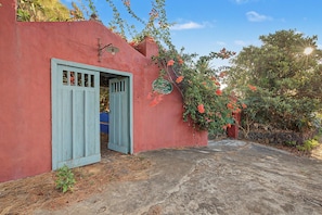 Un bâtiment rouge avec une porte bleue, un mur de pierre et un jardin où poussent des fleurs rouges.