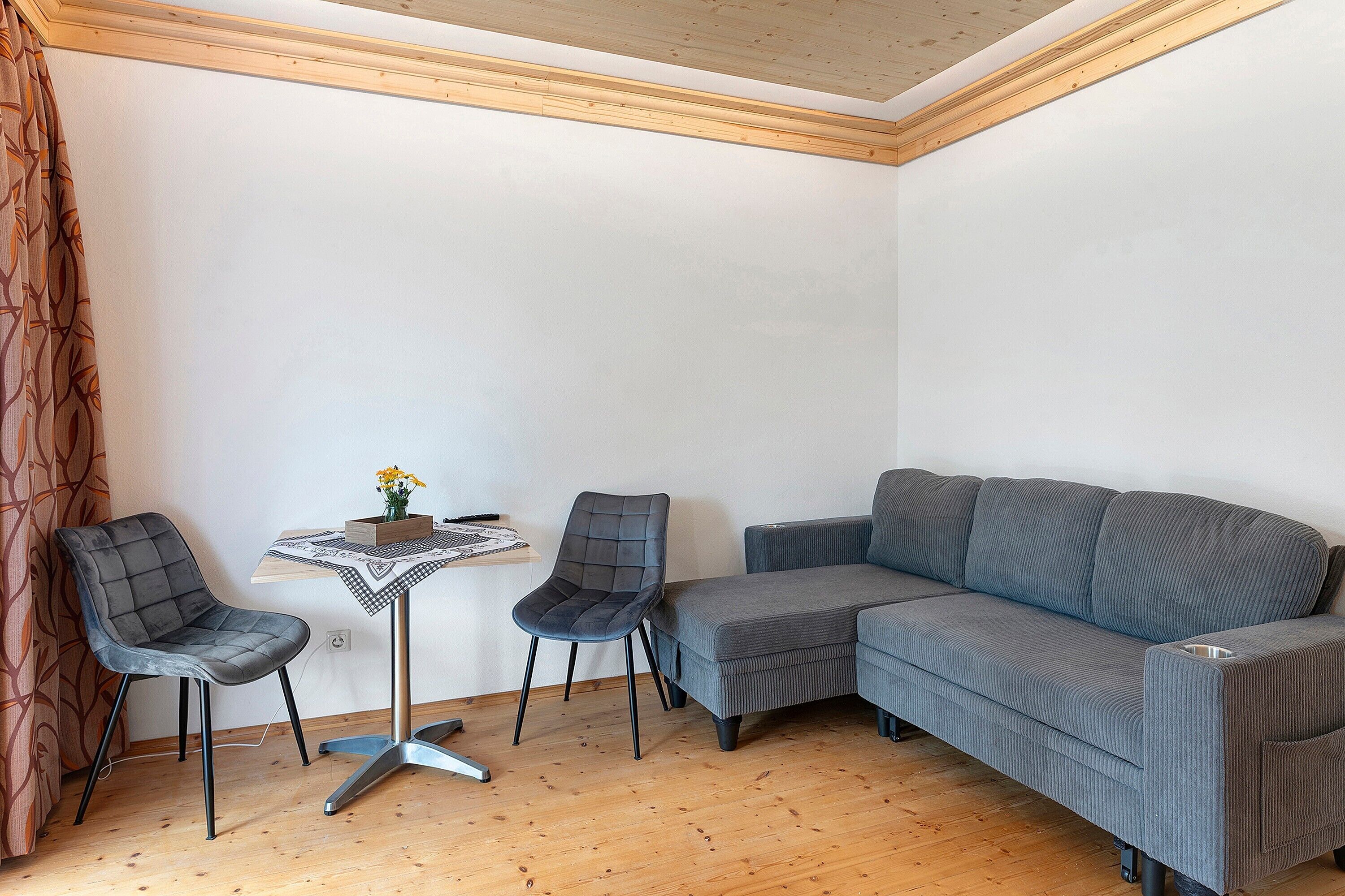 Ein Eckzimmer mit Holzboden, einem grauen Sofa, zwei Stühlen, einem kleinen Tisch mit Blumenvase und einer gemusterten Tischdecke.