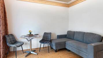 Ein Eckzimmer mit Holzboden, einem grauen Sofa, zwei Stühlen, einem kleinen Tisch mit Blumenvase und einer gemusterten Tischdecke.