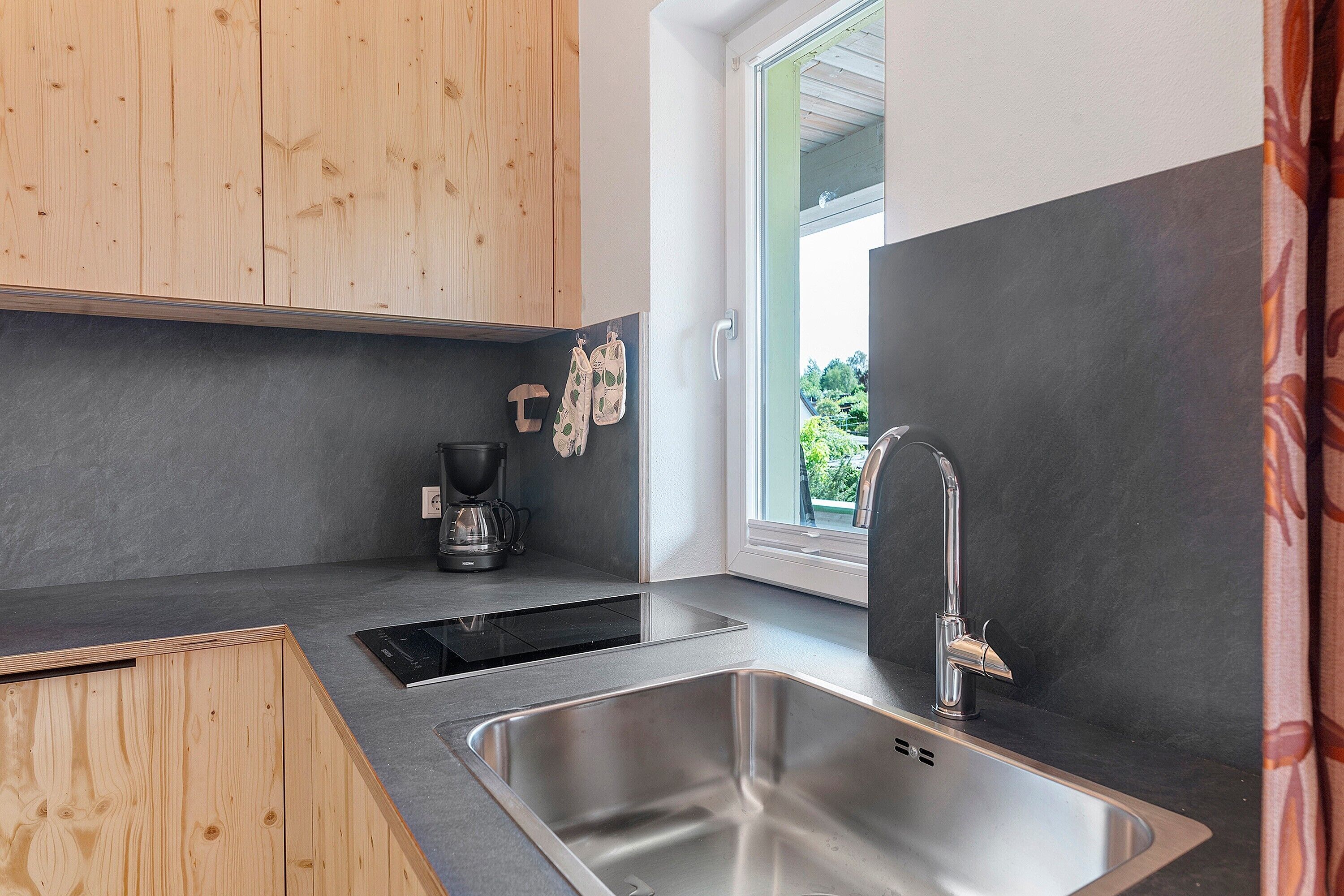 Eine moderne Küche mit Holzausbau, einer schwarzen Arbeitsplatte, einer Spüle aus Edelstahl und einem Fenster mit Blick auf Grünflächen.