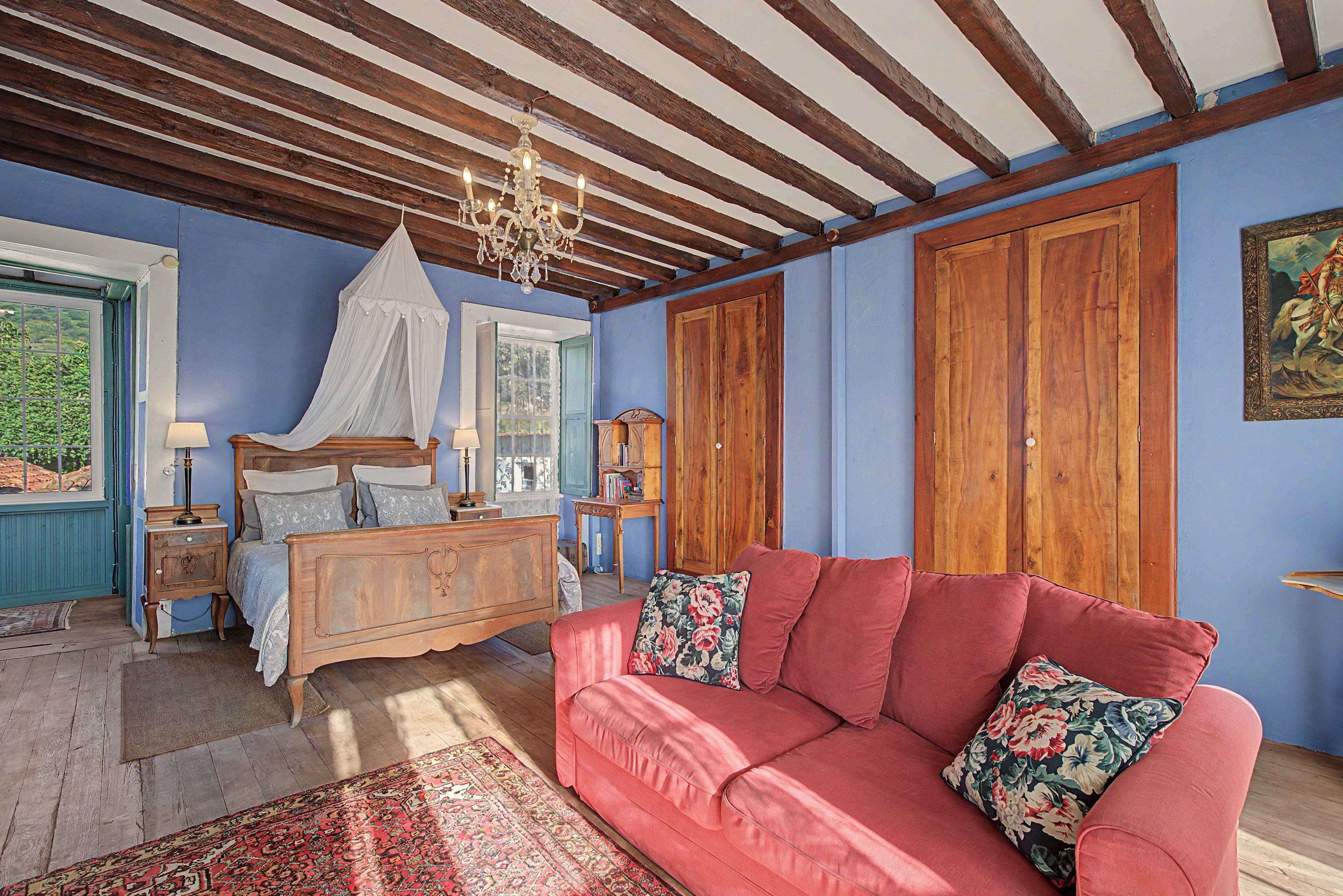 Quarto com cama, sofá vermelho, mobiliário de madeira, lustre e uma janela grande.