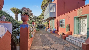Una estrecha calle adoquinada bordeada por edificios de paredes rojas y puertas verdes, que conduce a una zona cubierta con mesas al aire libre.