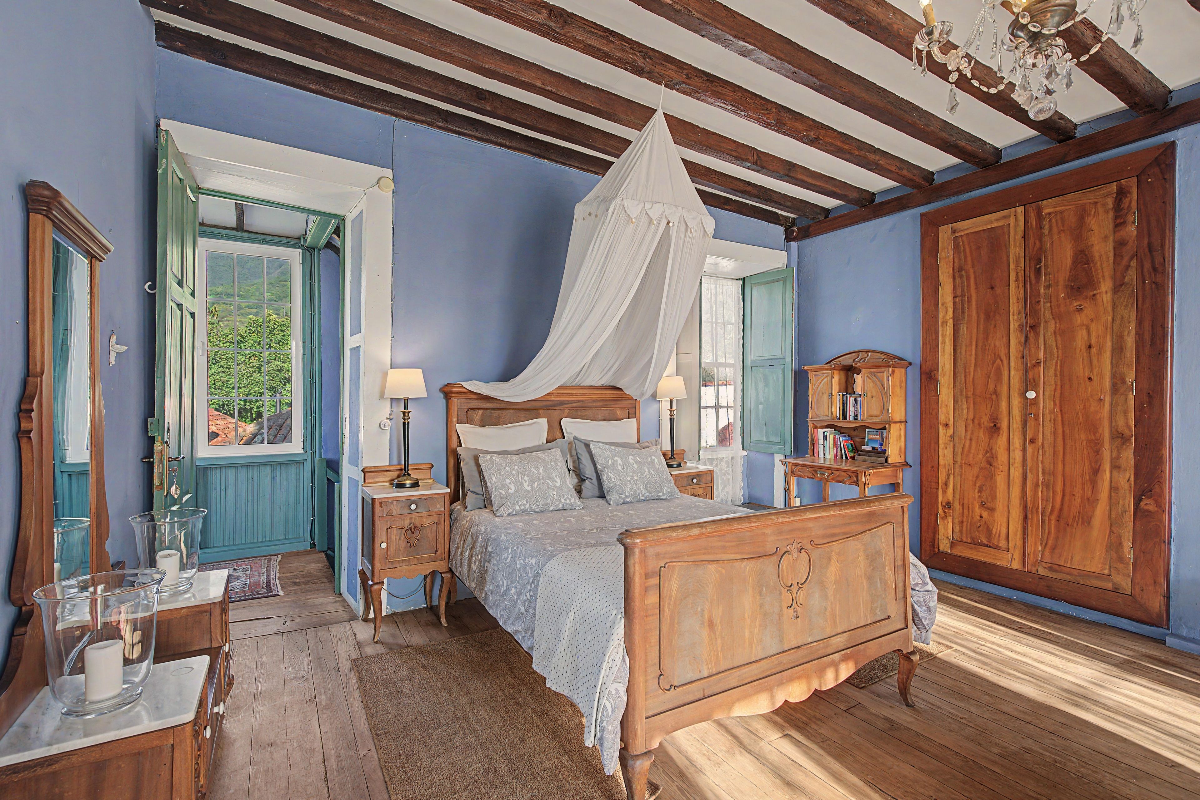 Quarto com cama de madeira, mesa de cabeceira, luminária, guarda-roupa e janela com vista para o verde.