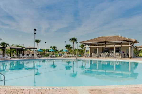 Una piscina con sillas de descanso y sombrillas, rodeada de palmeras y un pabellón cubierto.