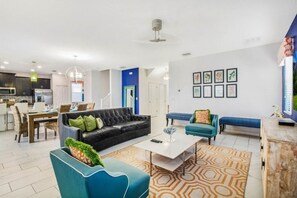Una sala moderna con un sofá de cuero negro, un sillón azul y una mesa de centro blanca. Se observa un comedor al fondo con una mesa y sillas.