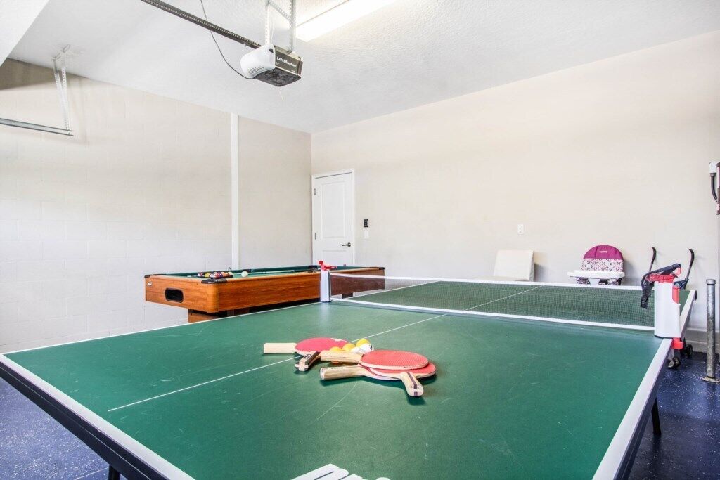 Une table de ping-pong avec raquettes et balles, une table de billard et un projecteur dans une pièce aux murs blancs.
