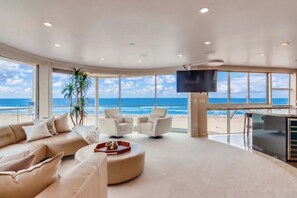 Un salón amplio con un gran ventanal con vistas a la playa, un televisor de pantalla plana montado en la pared y una moderna disposición de sofás.