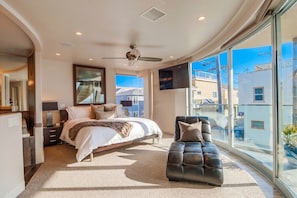 A modern bedroom with a bed, a seating area, a TV, and a large window offering a view of the outside. - Boardwalk Paradise 2 | New Mid-Stay Home w/ Endless Sunsets (San Diego)