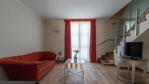 A modern living room with a red sofa, a small table with a vase, a TV on a stand, and a staircase with glass railings. - LOSTU Monforte (Monforte d'Alba)