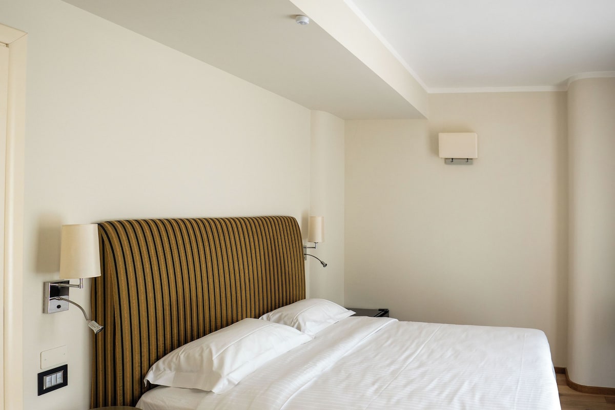 Ett hotellrum med en enkel säng, ett sängbord, en lampa och en vägglampa.