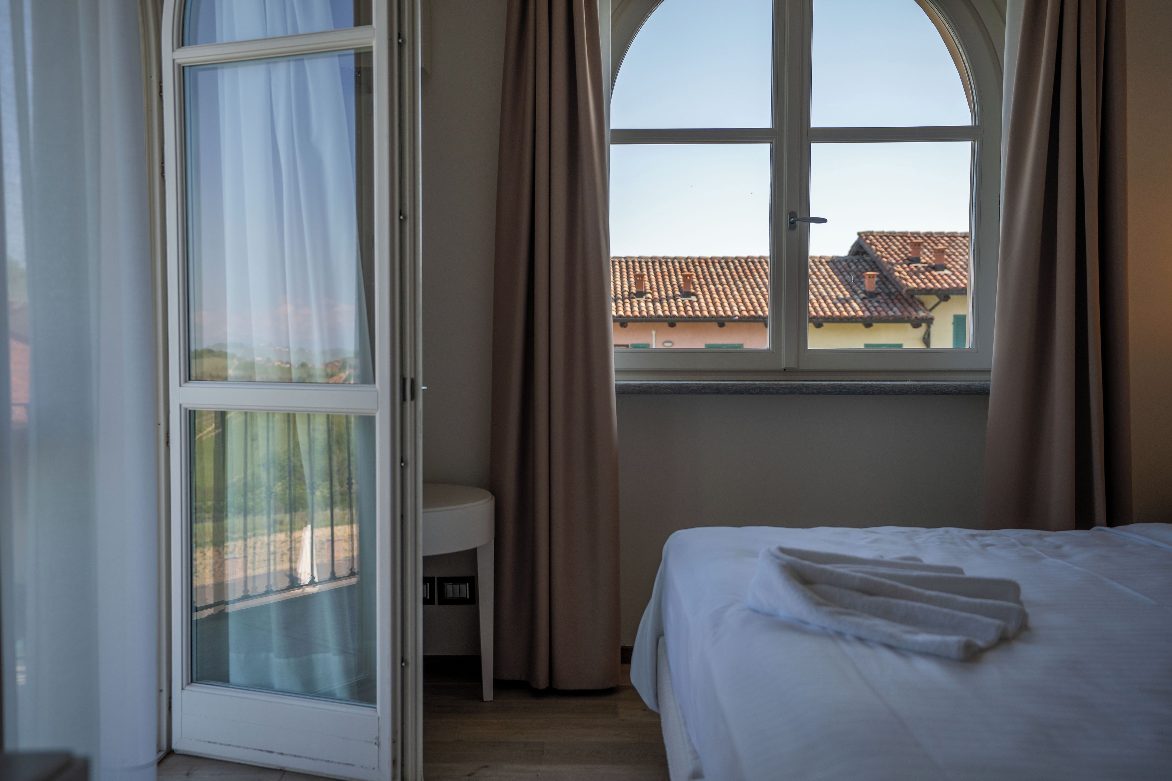 Ein Hotelzimmer mit Bett, Nachttisch, einem Fenster mit Vorhängen und Blick auf Gebäude.
