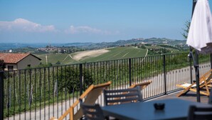 A terrace with a view of vineyards and distant buildings, featuring outdoor seating and a railing. - LOSTU Monforte (Monforte d'Alba)