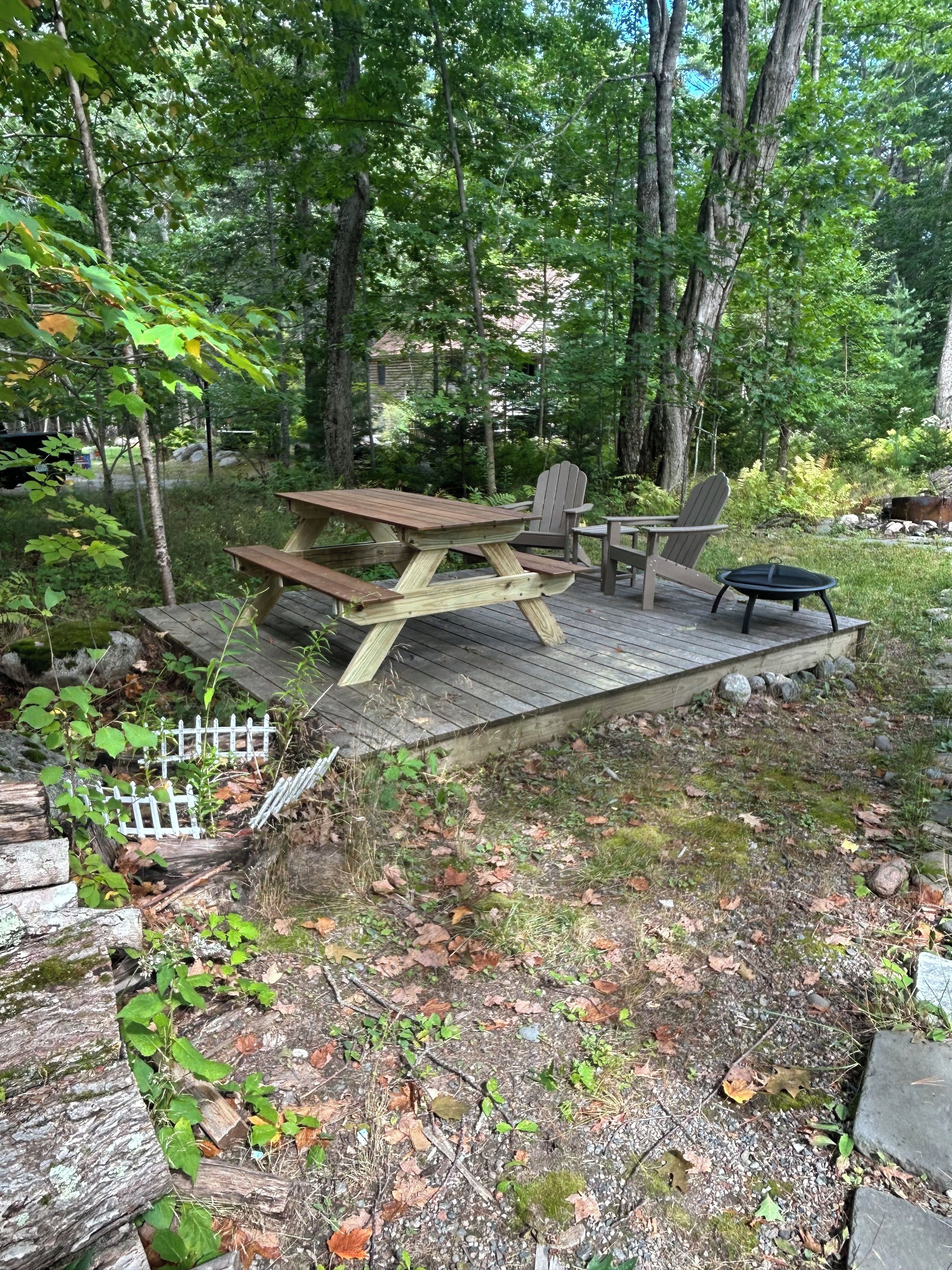 Eine Holzterrasse mit Picknicktischen und -stühlen, umgeben von Bäumen und einem kleinen Garten.