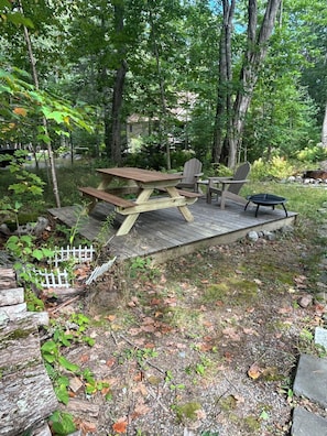 A wooden deck with picnic tables and chairs, surrounded by trees and a small garden.