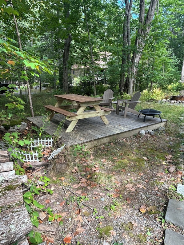 A wooden deck with picnic tables and chairs, surrounded by trees and a small garden.