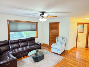 A living room with a leather sofa, a glass coffee table, a patterned armchair, a wooden floor, and a ceiling fan.