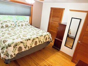 Ein Schlafzimmer mit einem Bett, einem Holzschrank, einem Spiegel und einem Fenster mit Jalousien.